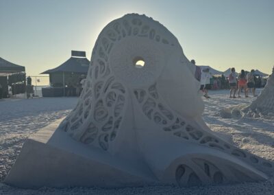 Abstract sand sculpture with sunlit backlighting, surrounded by tents and festival visitors.