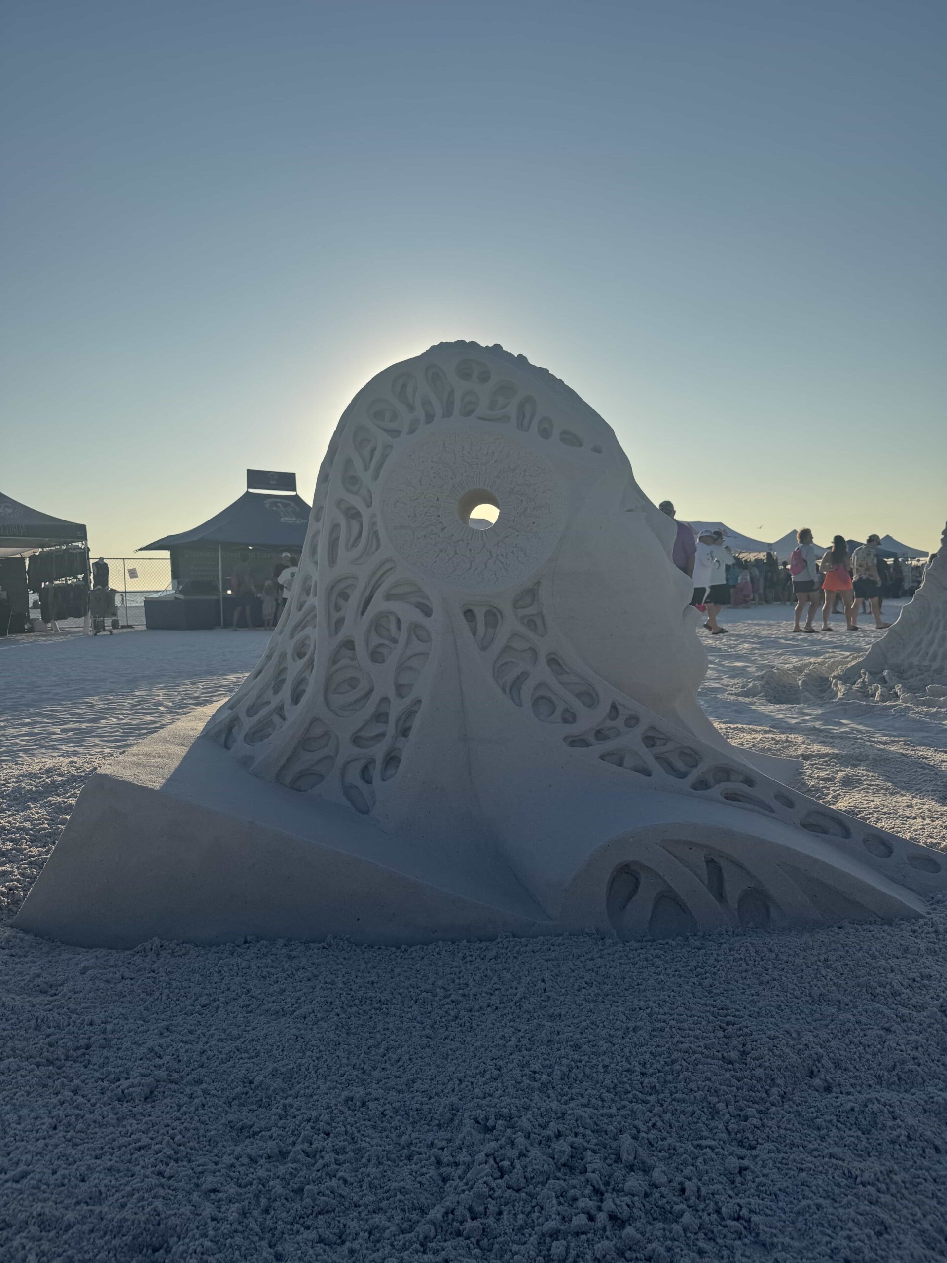 Abstract sand sculpture with sunlit backlighting, surrounded by tents and festival visitors.
