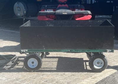 Black utility cart near red ATV and trailer at outdoor loading area with coiled hose on ground.
