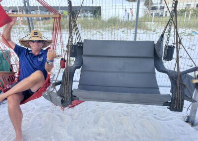 Man in straw hat on red beach swing, smiling beside empty woven bench swing on sandy beach.