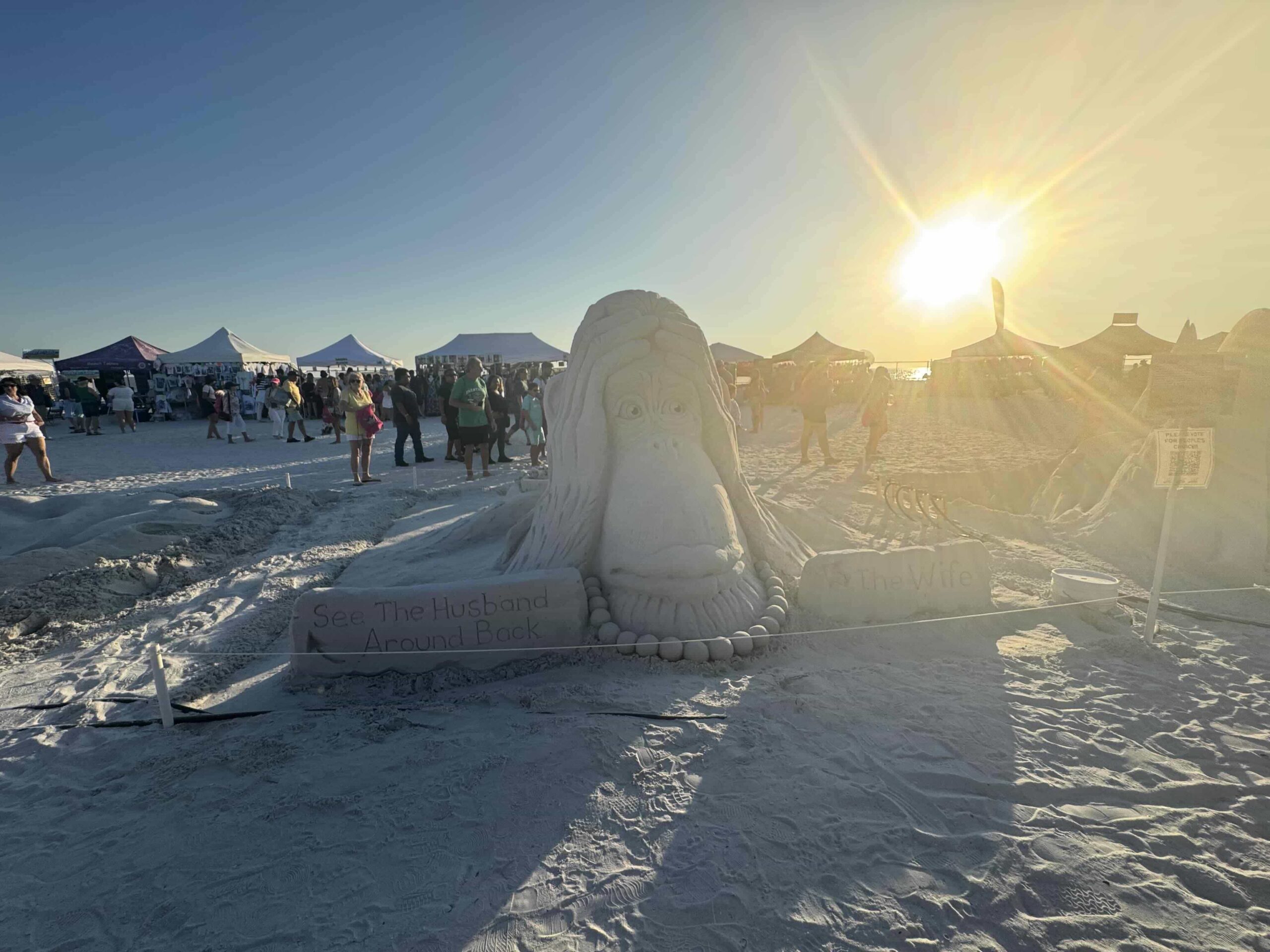 Sand sculpture of a monkey's face labeled "The Wife," with sunset glow and event crowd in background.