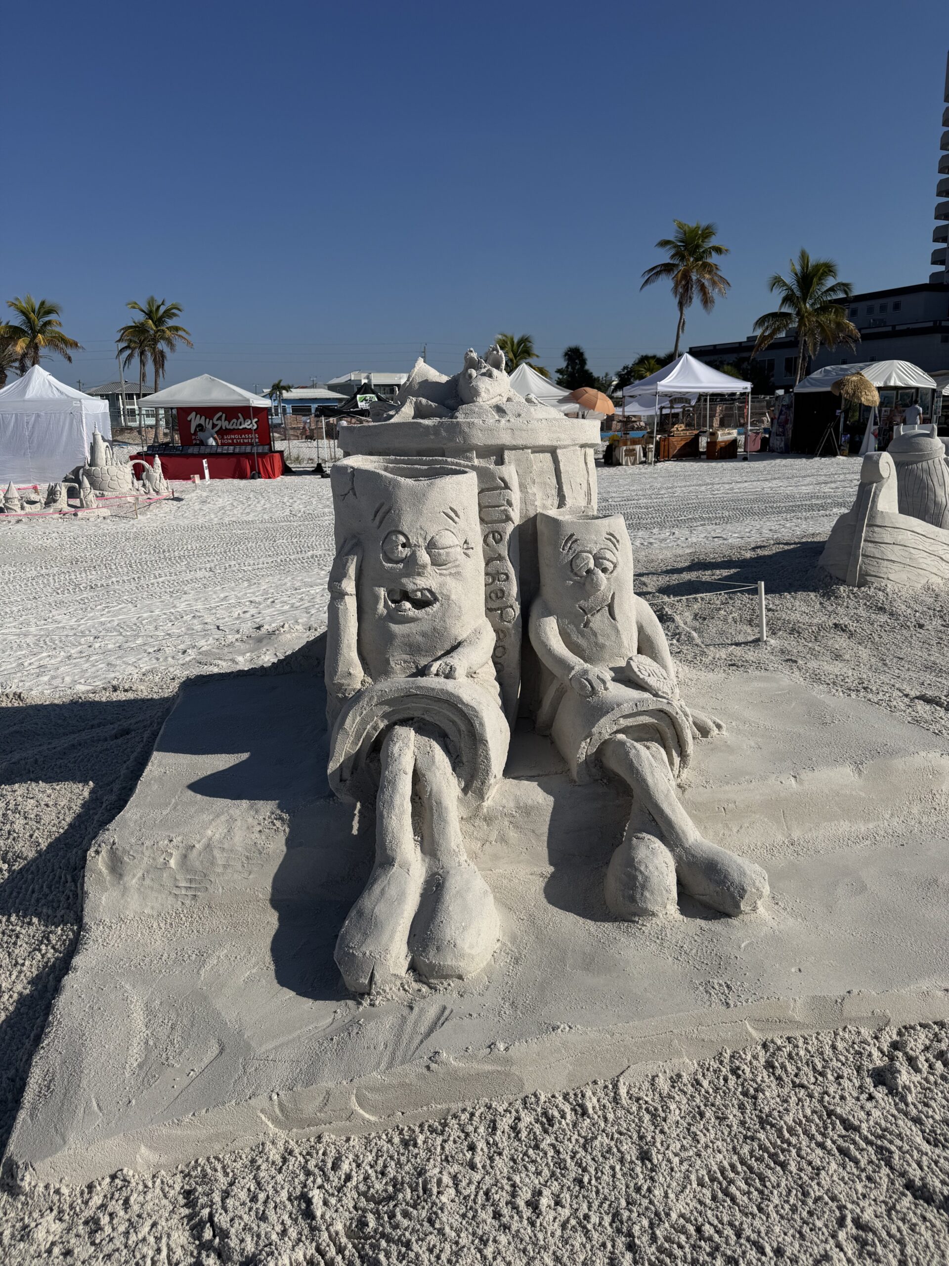 Sand sculpture of SpongeBob and Patrick relaxing in beach chairs at a sunny outdoor event.