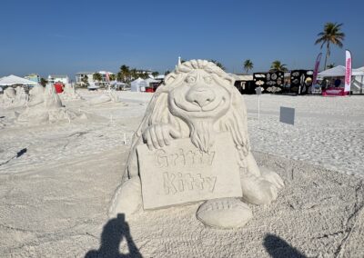 Whimsical sand character holding a “Gritty Kitty” sign, with other sculptures and tents nearby.
