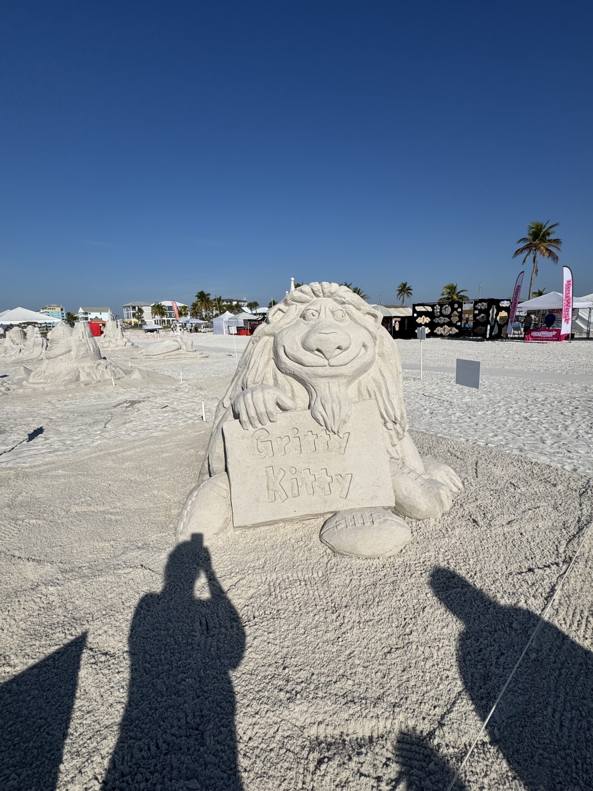 Whimsical sand character holding a “Gritty Kitty” sign, with other sculptures and tents nearby.