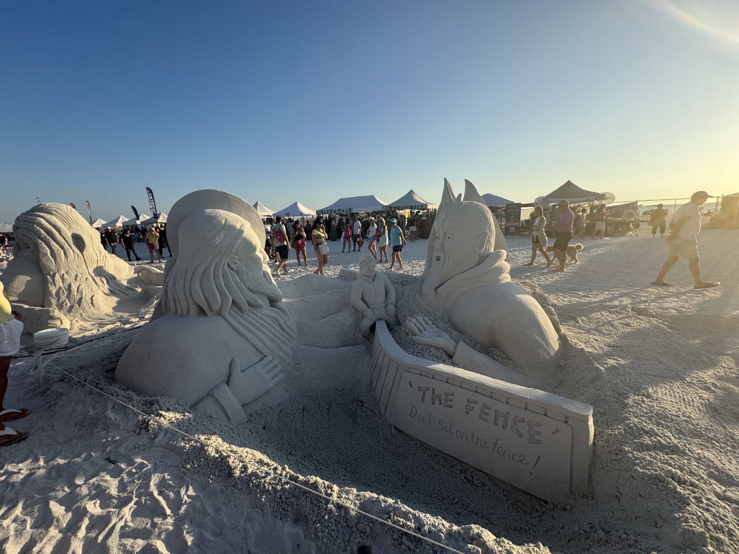 Two sand figures divided by a fence sculpture with the message “Don’t sit on the fence!”