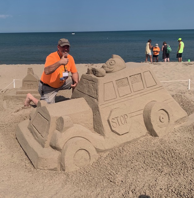 A man in an orange shirt and cap kneels beside a large sand sculpture of a school bus, giving a thumbs-up. The sculpture features carved wheels, windows, a “STOP” sign, and a cartoon-like frog character perched on top. Beachgoers and the ocean under a clear blue sky complete the festive scene.