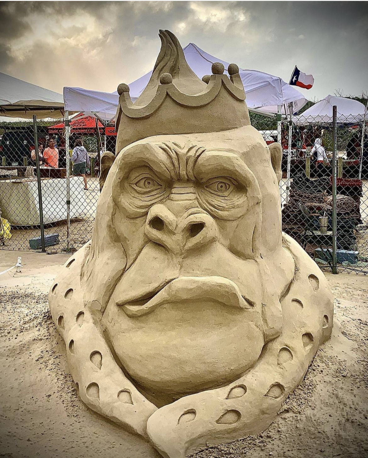 A detailed sand sculpture of a gorilla dressed in regal attire, wearing a crown and a spotted robe. The gorilla’s stern expression enhances its commanding presence. The sculpture is part of an outdoor competition or festival, with tents, fencing, and people nearby. A Texas state flag is visible in the background.