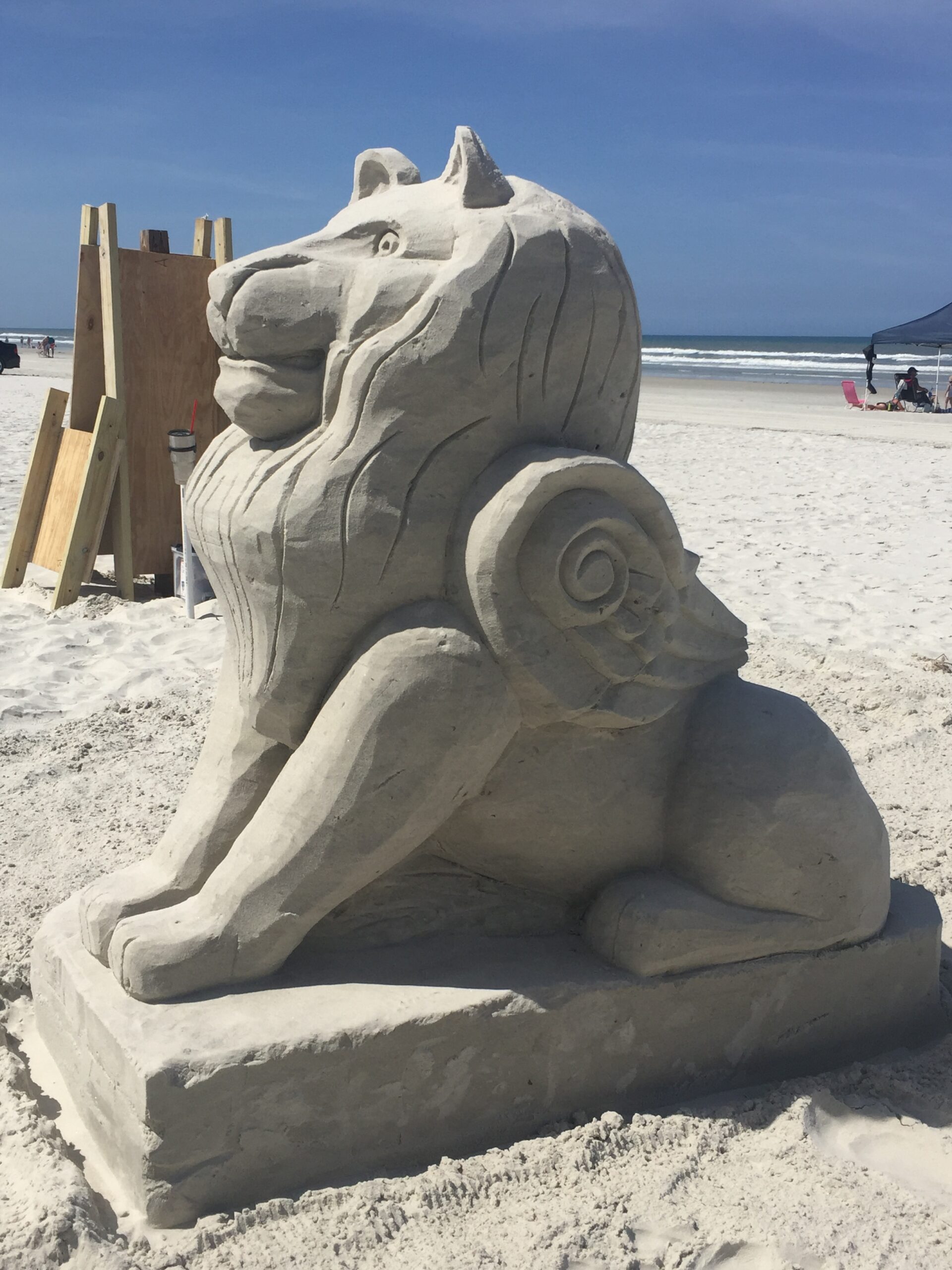 A detailed sand sculpture of a lion with carved mane and spiral-shaped wings sits on white beach sand. The ocean and clear blue sky form a serene backdrop, with wooden boards and a container nearby hinting at the sculpting process.