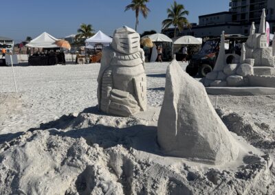 Sand sculptures of knight, rock, and castle at beach fest with tents and palm trees.