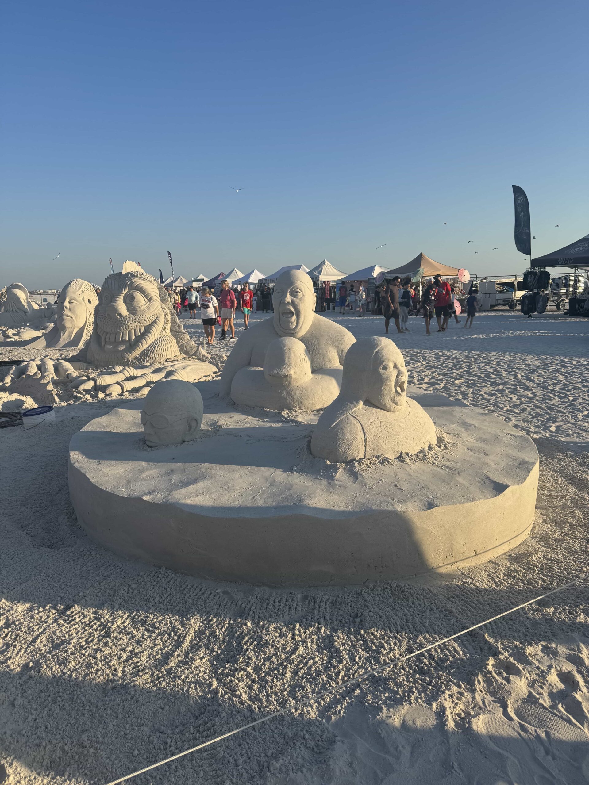 Four expressive sand heads in a circular base, with other sculptures and festival tents in the background.