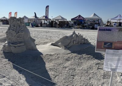 Sand sculpture of ocean-themed face with coral and shell at beach event, artist sign in foreground.