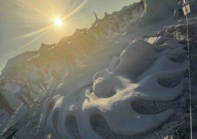 Large surreal sand sculpture at sunset beach fest, with crowd and tents in background.