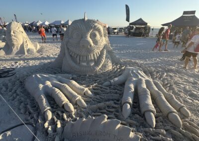 Sand creature with claws and grin titled “Rising from the Deep” at beach sculpture event.