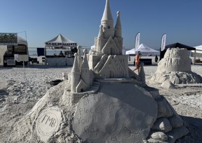 Sandcastle with “Oh the Places You'll Go!” plaque at sunny beach fest, tents in background.