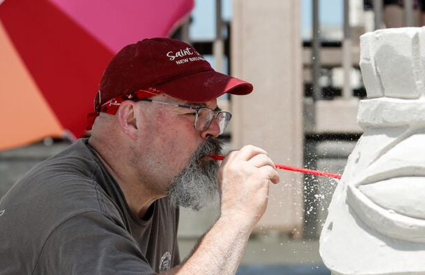 A sand sculptor wearing a red cap, glasses, and dark shirt uses a red straw to blow away fine sand from a detailed sculpture resembling a stylized face or mask. Behind them, a large red and pink umbrella and wooden boardwalk structure add to the beachside setting.