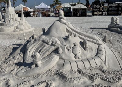 Sand sculpture of volcano, bear surfing, and fruit at beach event with palm trees and tents.