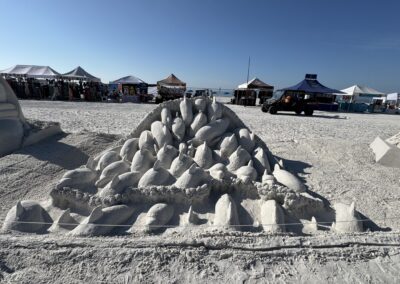 Sand sculpture of dolphins stacked in a pyramid, with tents and beachgoers in the background.