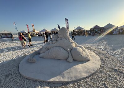 Sand sculpture of woman with cats titled “Purrfect Love” at beachside art event with vendor tents.