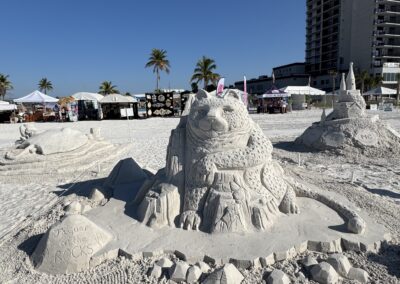 Sand jaguar perched on rocks with “Jaguar on the ROCKS” plaque at beach sculpture event.