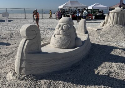 Sand Viking with horned helmet seated in carved boat at beach fest with ocean in background.