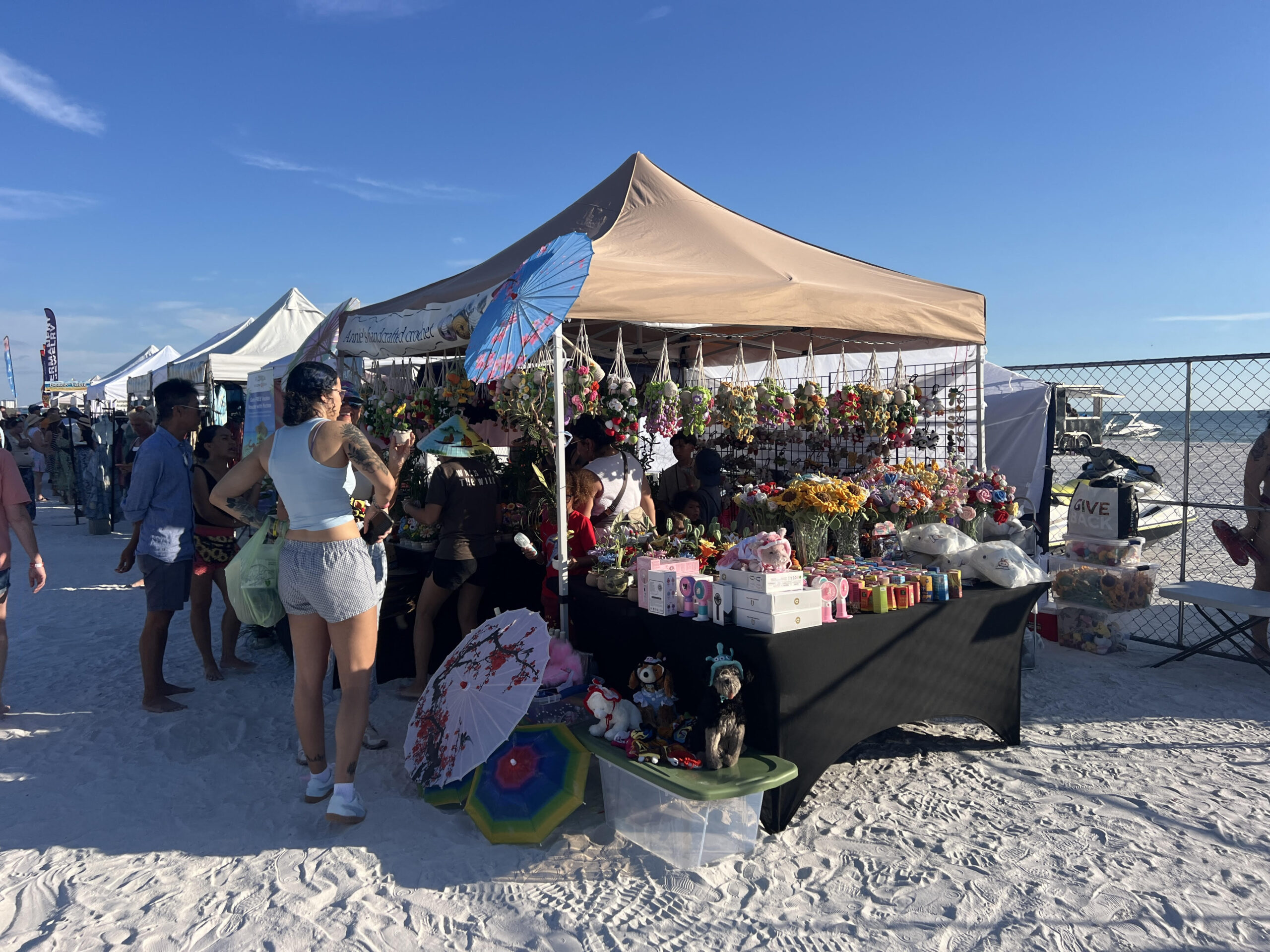 unnamed Beach market tent with plush toys, flowers, and “Anna’s Handcrafted Creations” sign, jet ski in background.
