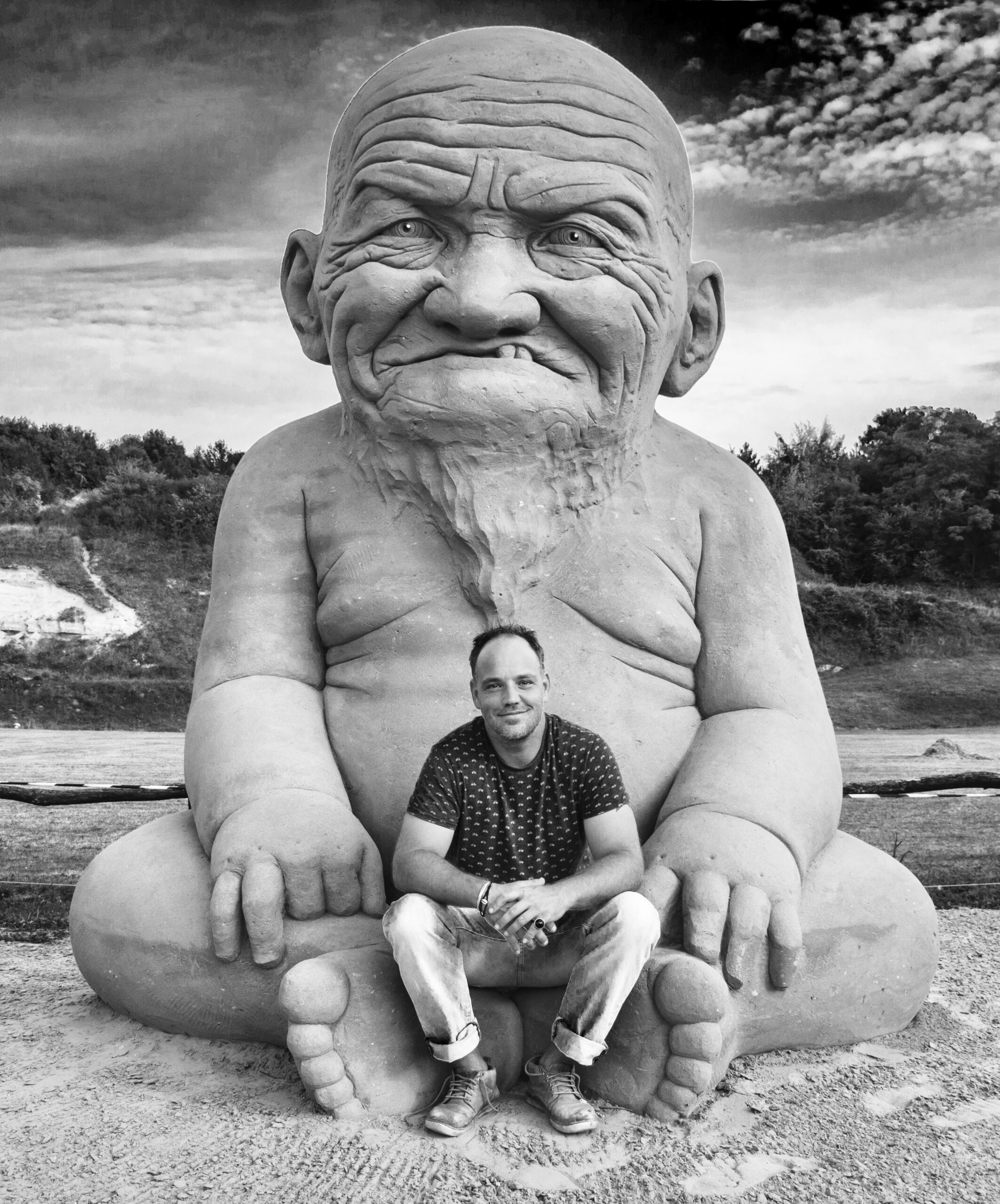 Man mimics pose of giant bearded sand figure, seated between its legs at outdoor event.