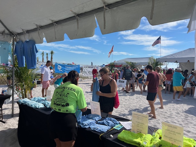 Volunteers in neon shirts assist attendees under a large white beach tent with sponsor banners behind them.